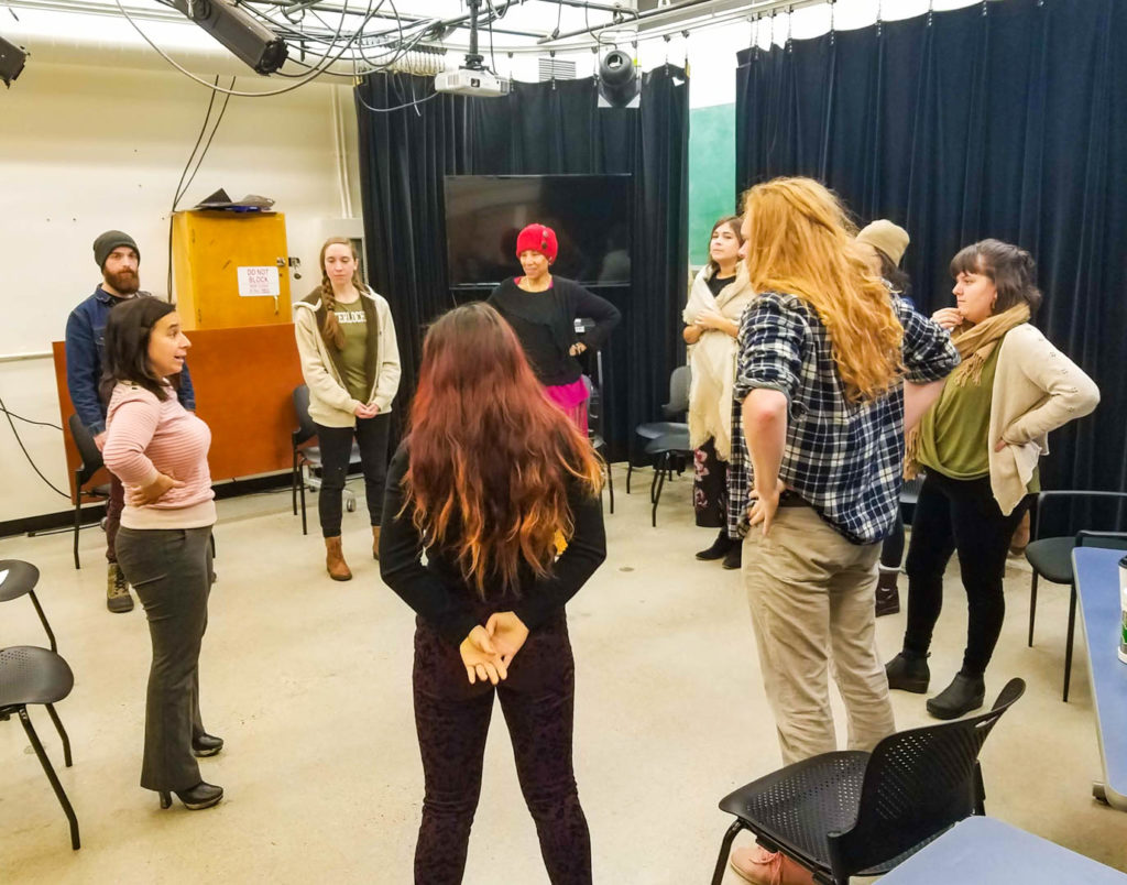 Students in a theatre classroom standing in a circle listening to Erica, at left, hands on her hips.